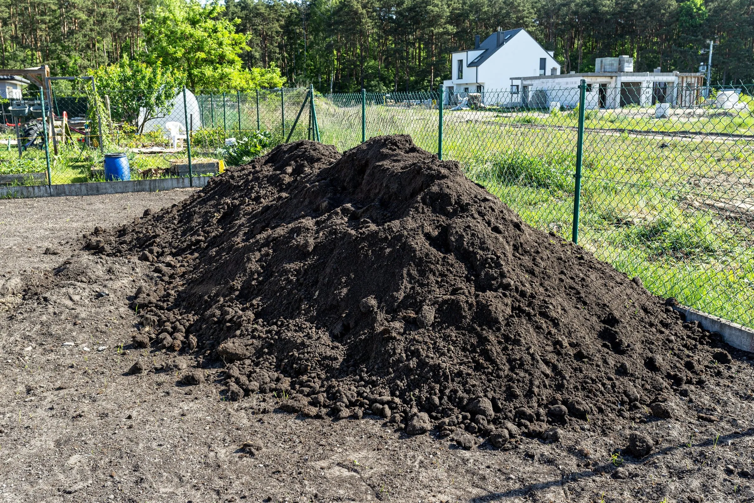 A large pile of dark excavated soil sits at a construction site in front of a wire fence and forest.