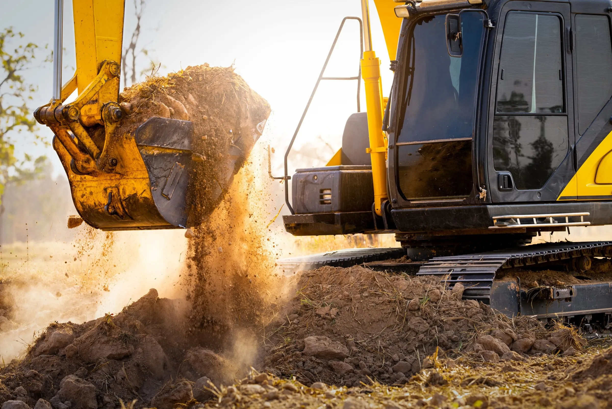 Close-up of a yellow excavator bucket dumping soil at sunset.