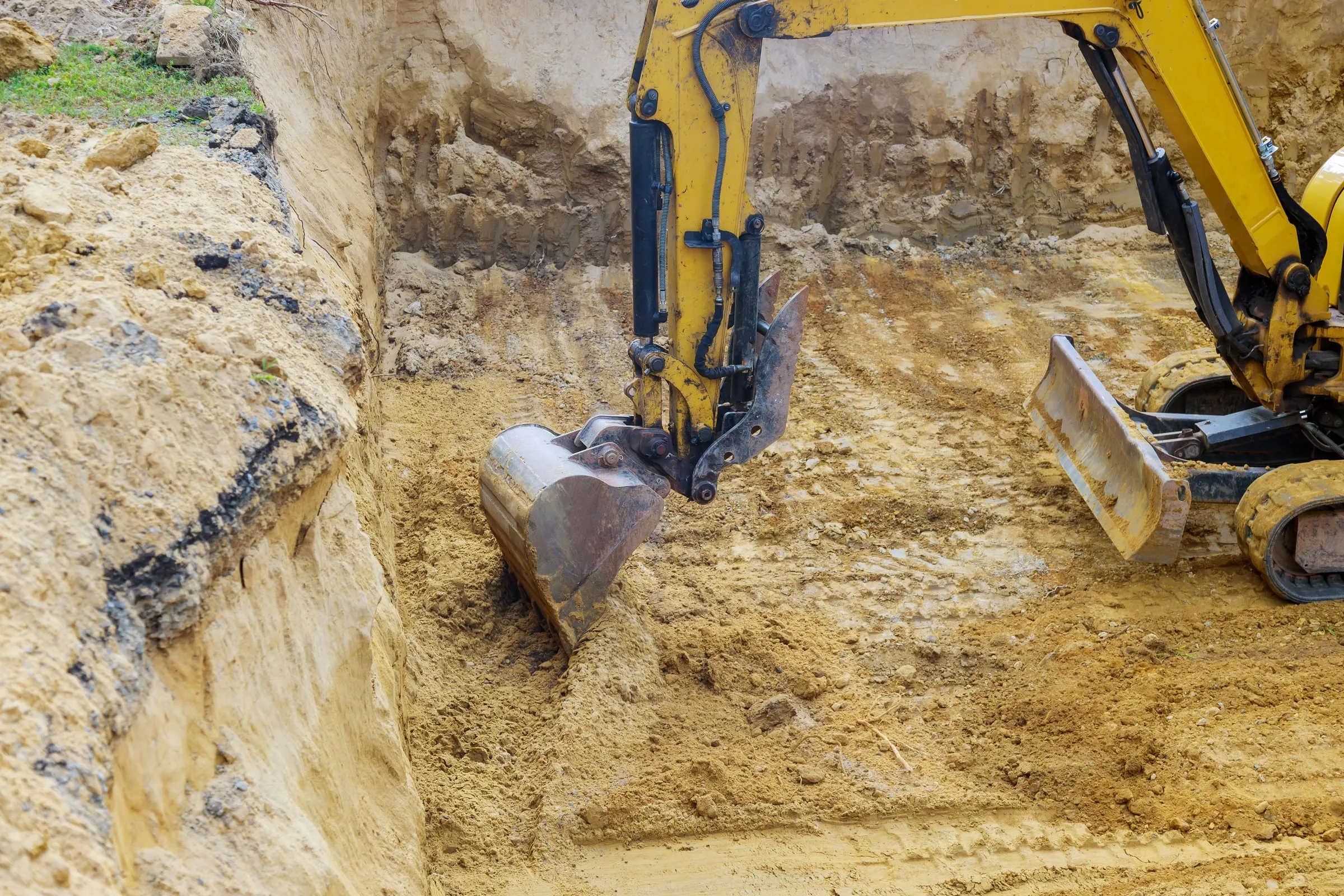 Close-up of a yellow excavator bucket digging into dark soil to form a foundation pit at a residential construction site.