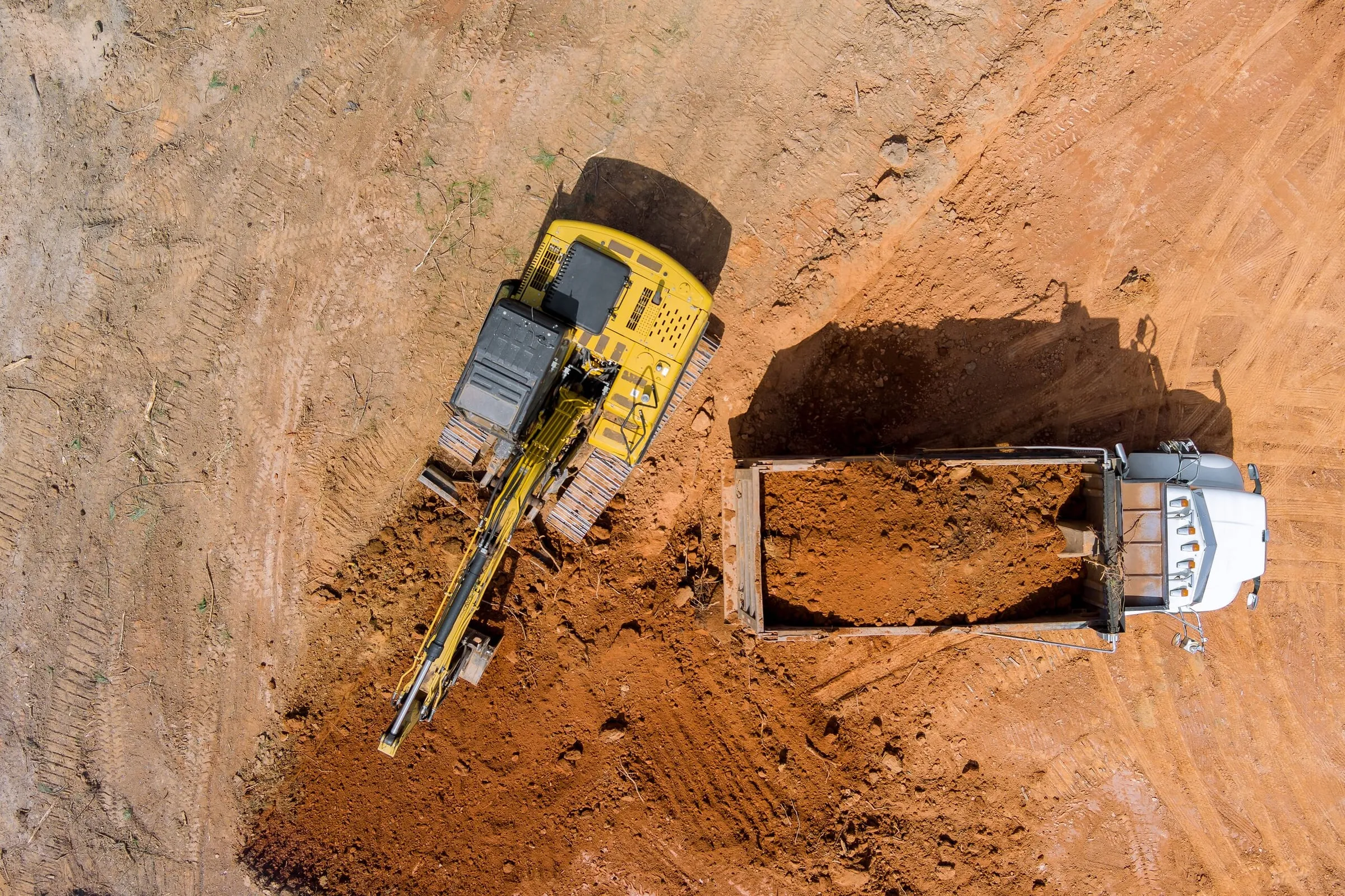Aerial view of a yellow excavator loading red dirt into a white dump truck.