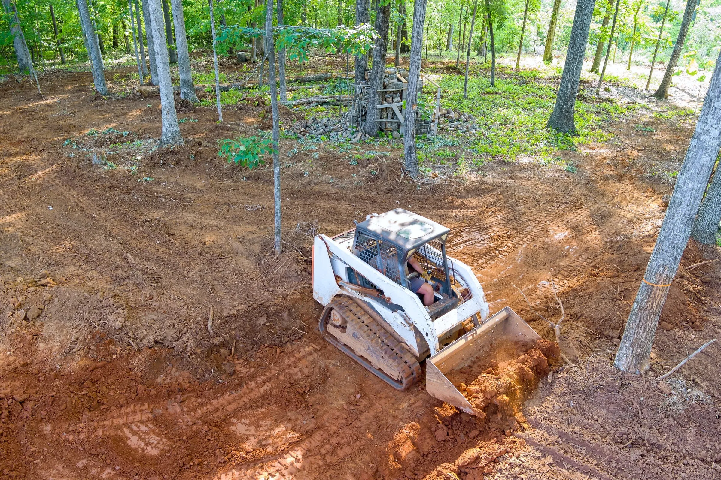 Aerial view of a white skid steer loader grading and clearing soil in a wooded area.